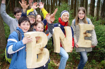 Kinder stehen im Wald und halten aus Holz ausgesägte Buchstaben B, N und E