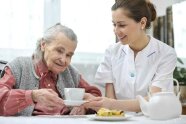 Frau reicht einer Seniorin Tasse auf Untertasse, davor auf dem Tisch Kuchenstück und Kaffeekanne 