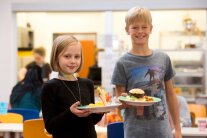 2 Kinder stehen in Kantine nebeneinander und halten einen Teller mit Speisen 