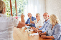 Referentin steht vor vier Senioren, die am Tisch nebeneinander sitzen 