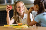 Mädchen mit Apfel in der Hand unterhält sich mit anderem Mädchen am Essenstisch.